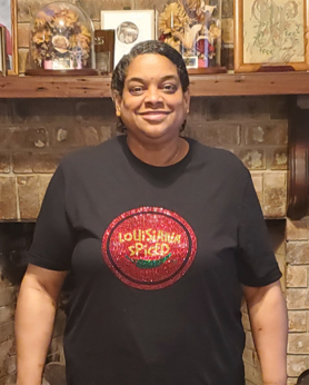 Person wearing a black Louisiana Spiced t-shirt with a red logo in front of a brick wall.